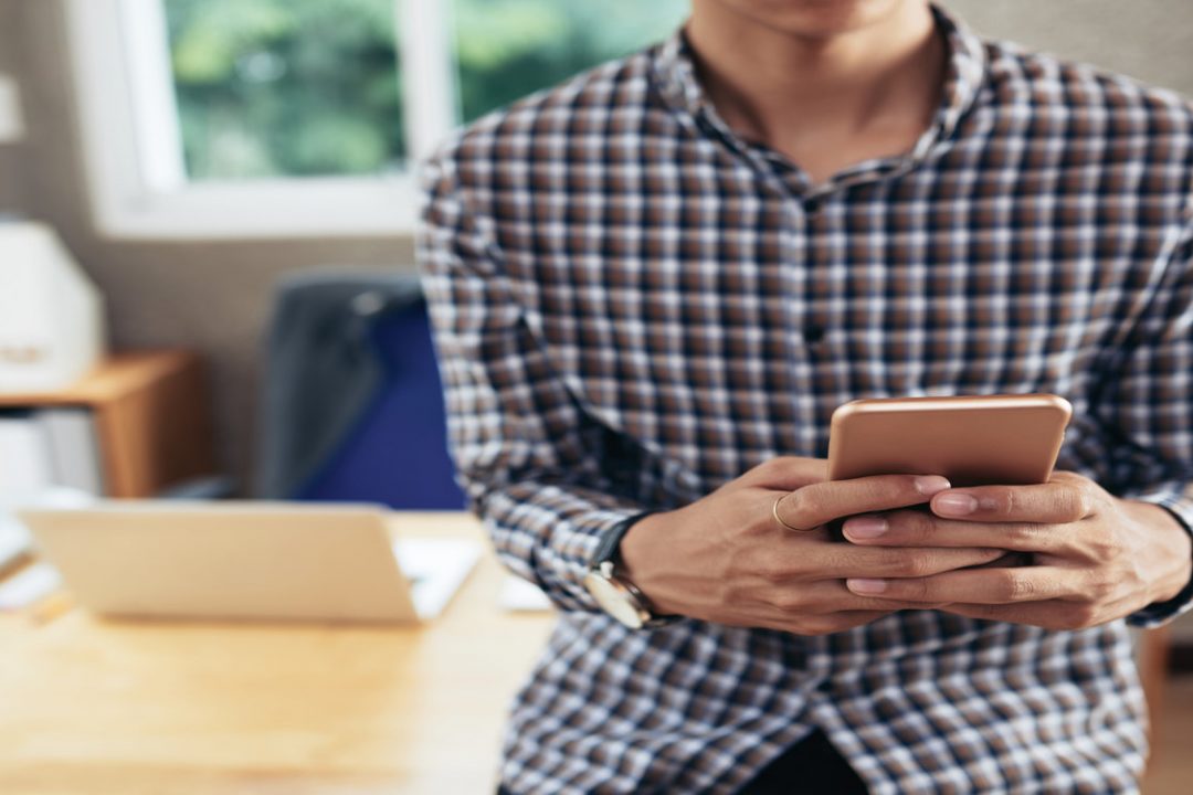 Crop shot of unrecognizable man sitting on office desk and texting on smartphone on blurred background